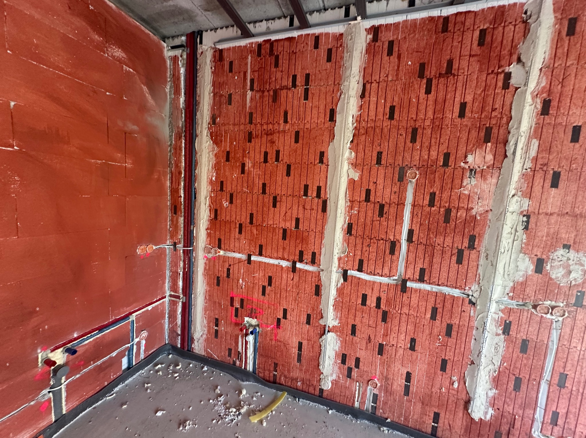 Czerwona ściana z instalacją elektryczną i rurami, częściowo pokryta zaprawą. Widoczne otwory w cegłach i szary betonowy sufit. Wnętrze w trakcie prac wykończeniowych.