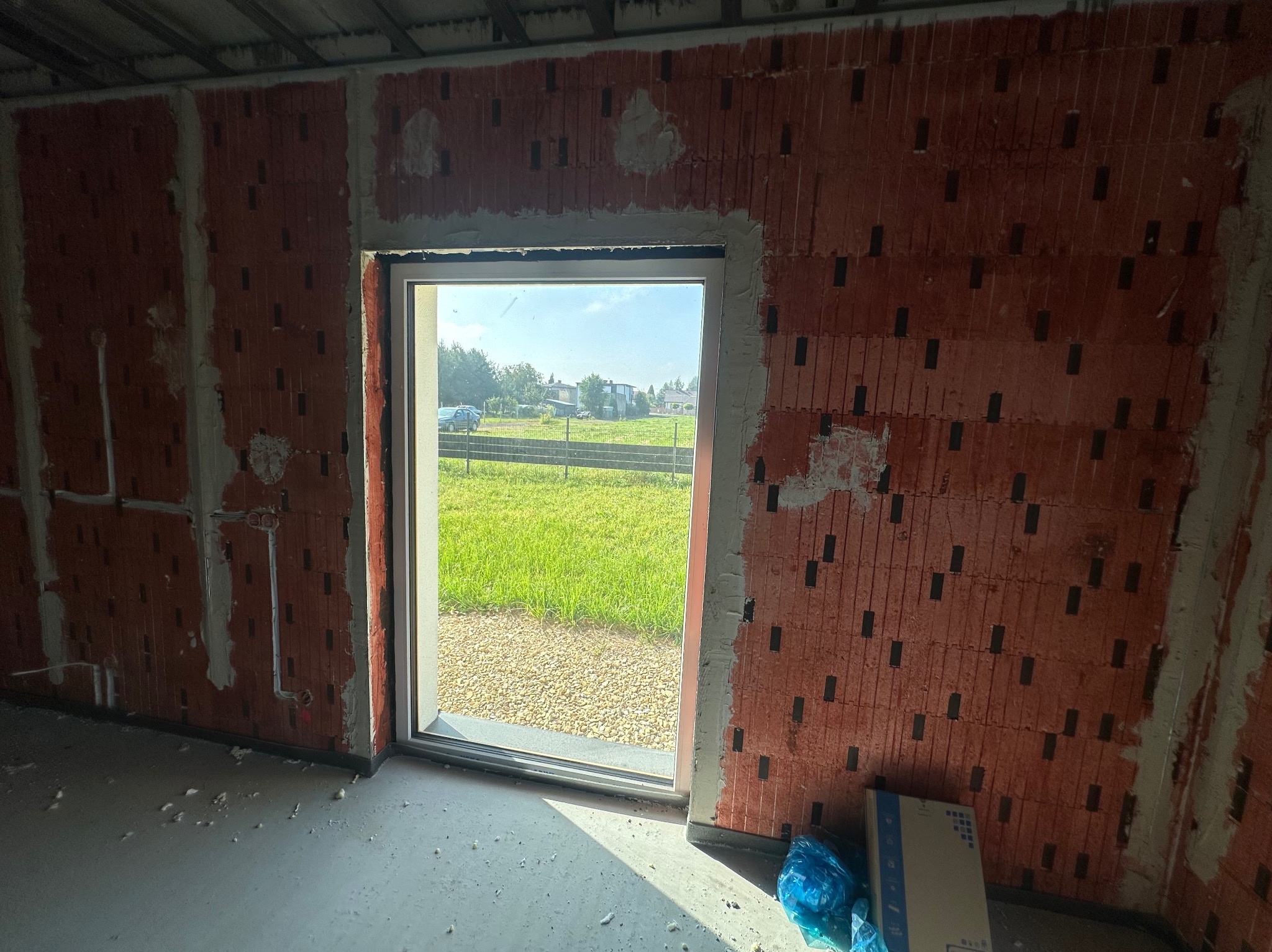 Surowy stan wnętrza z oknem na zewnątrz. Czerwone cegły z widocznymi spoinami, surowy beton na podłodze, instalacje na ścianach. Widok na zieleń i domy.