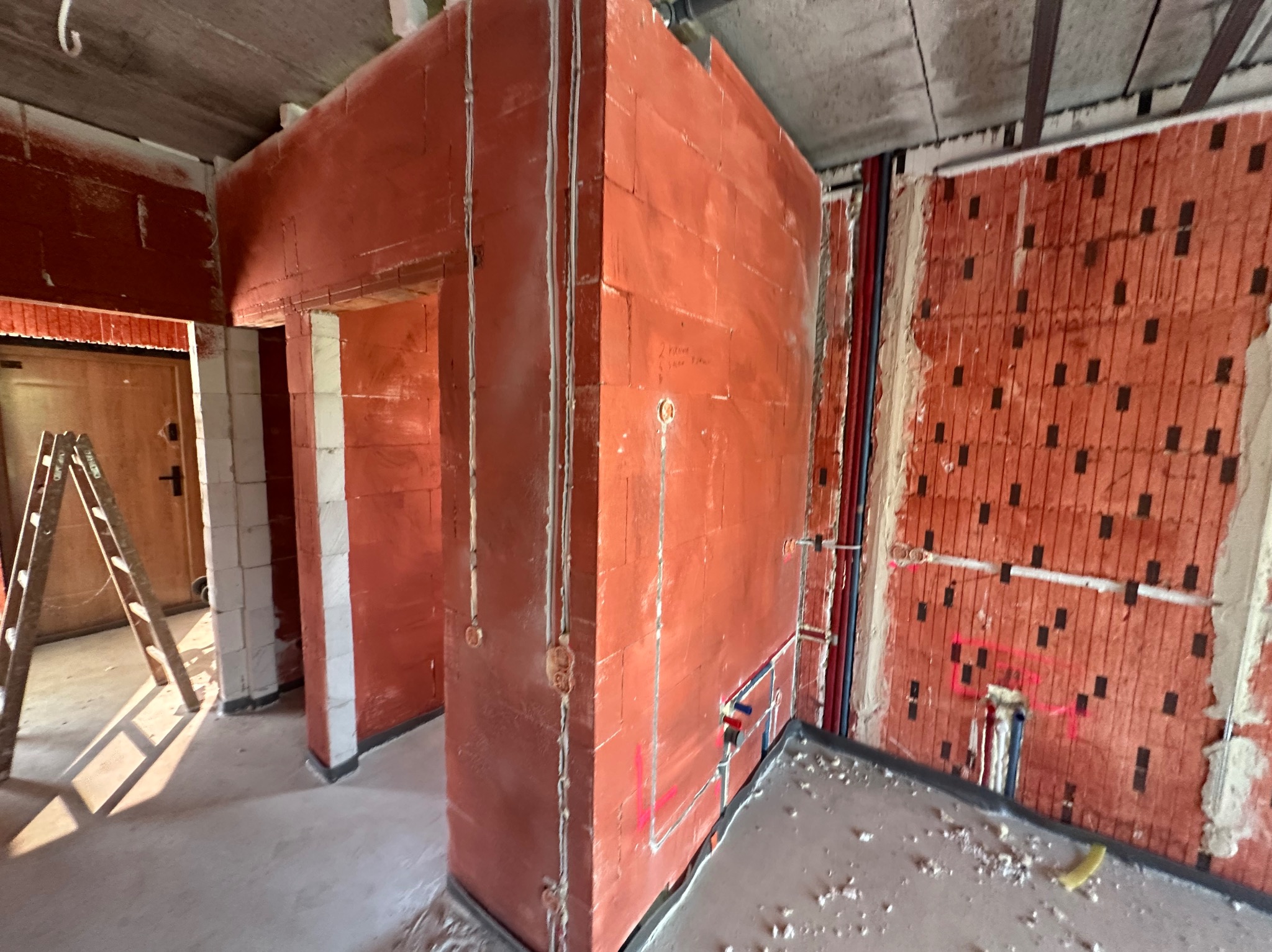 Wnętrze w trakcie budowy: ściany z czerwonej cegły z instalacjami elektrycznymi i hydraulicznymi. Widoczna drabina i surowy betonowy strop.