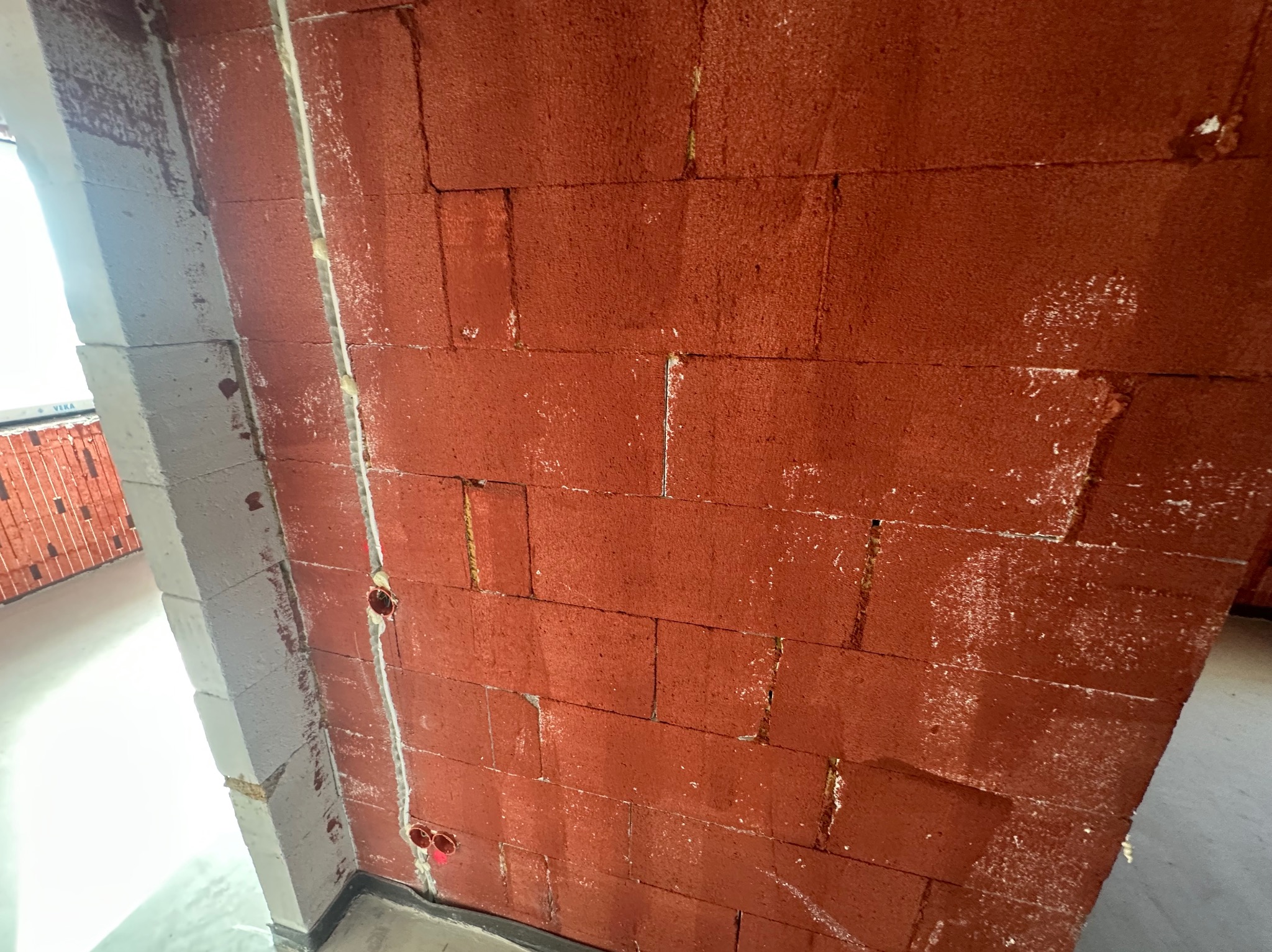 Czerwona ściana z pustaków ceramicznych z widocznymi otworami na instalacje elektryczne i fragmentem ościeżnicy okiennej w trakcie prac wykończeniowych.