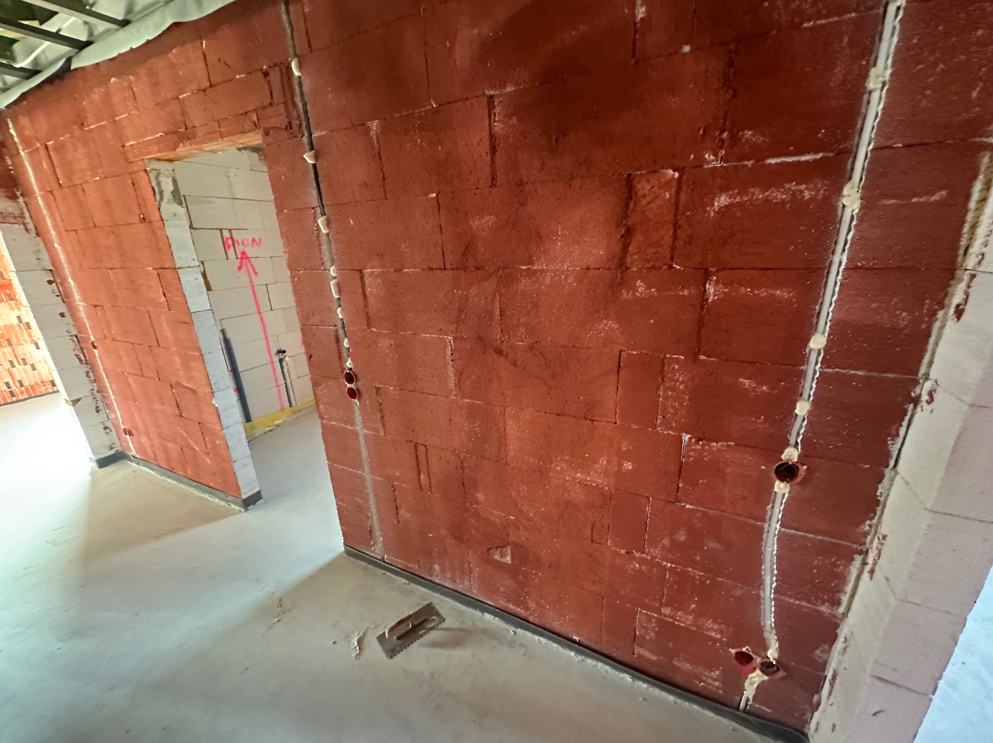 Czerwona ściana z bloków, częściowo otynkowana, z widoczną instalacją elektryczną i narzędziem na podłodze. W tle widoczne surowe ściany z białych bloków.