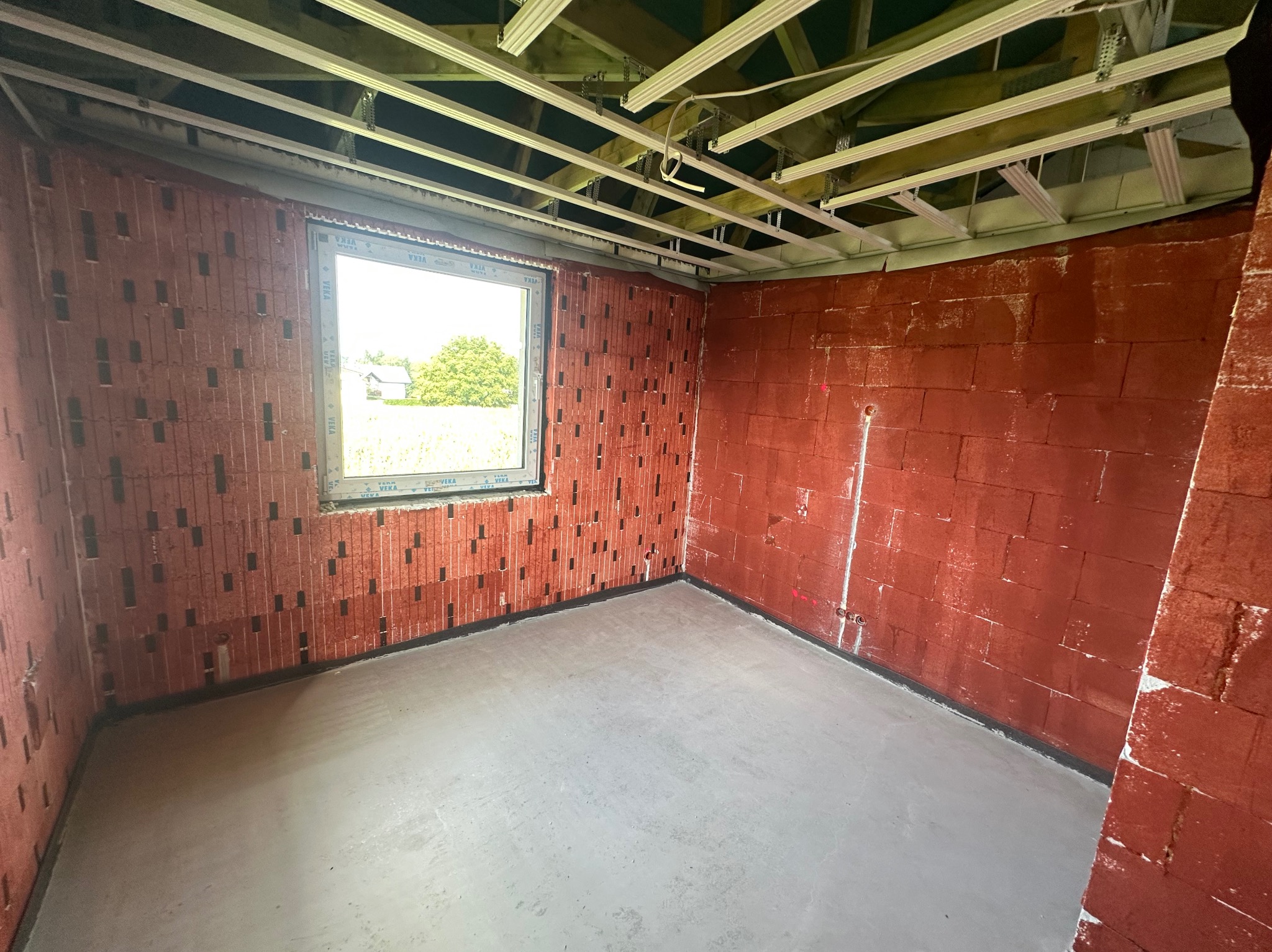 Surowy pokój z czerwonej cegły z oknem i wylewką, widoczne profile sufitowe. Świeża wylewka podłogowa w nowym budynku. Widok na zewnątrz przez okno.