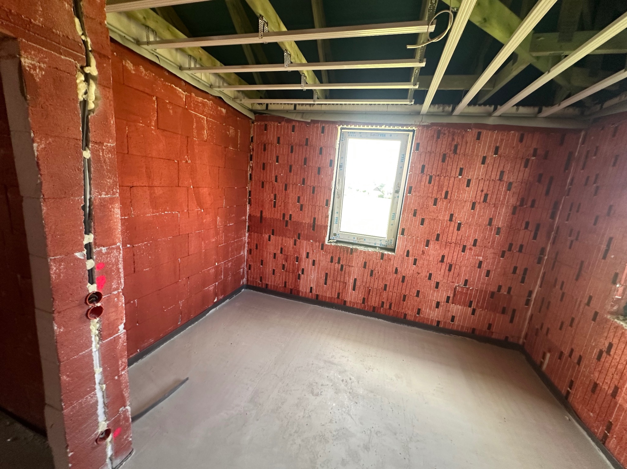 Surowy stan wnętrza: czerwona cegła dziurawka, wylewka podłogowa, okno z PCV i metalowy stelaż sufitowy. Widoczne instalacje elektryczne w ścianie.