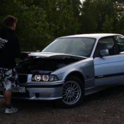 Mężczyzna w bluzie z kapturem i wzorzystych szortach stoi przy srebrnym BMW E36 z otwartą maską, widoczne okrągłe światła ringowe i otwarte drzwi kierowcy.