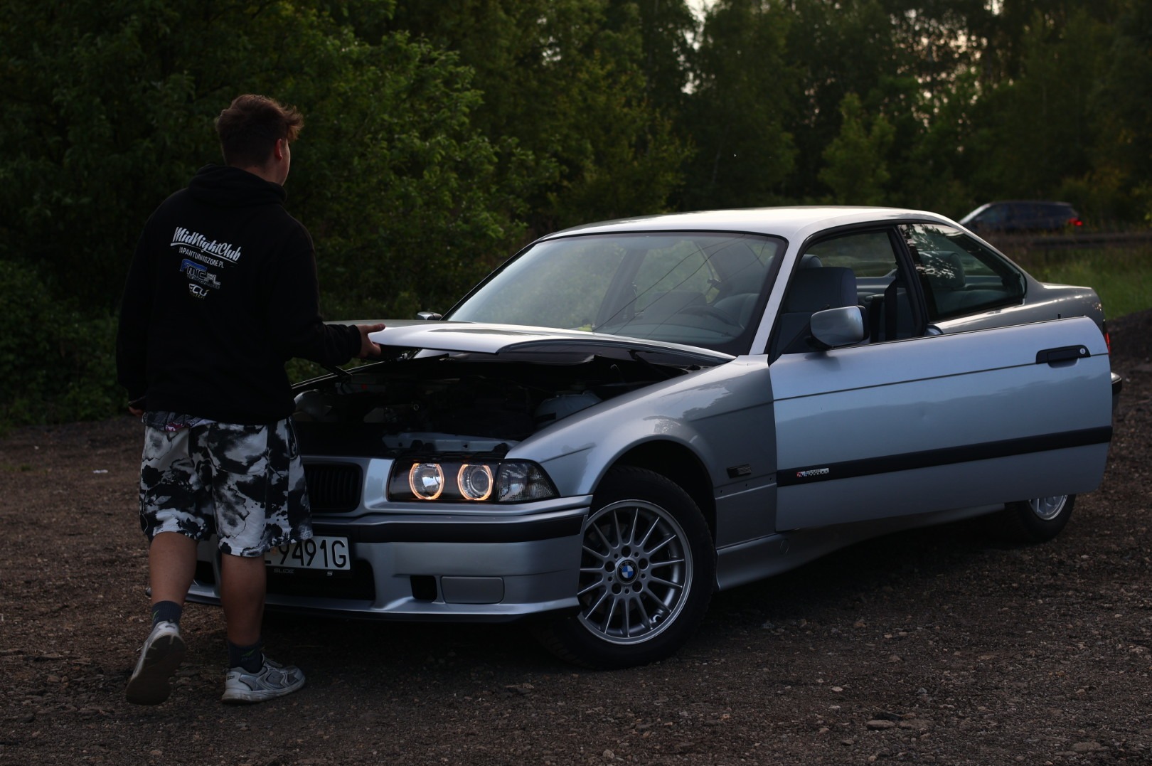 Mężczyzna w bluzie z kapturem i wzorzystych szortach stoi przy srebrnym BMW E36 z otwartą maską, widoczne okrągłe światła ringowe i otwarte drzwi kierowcy.