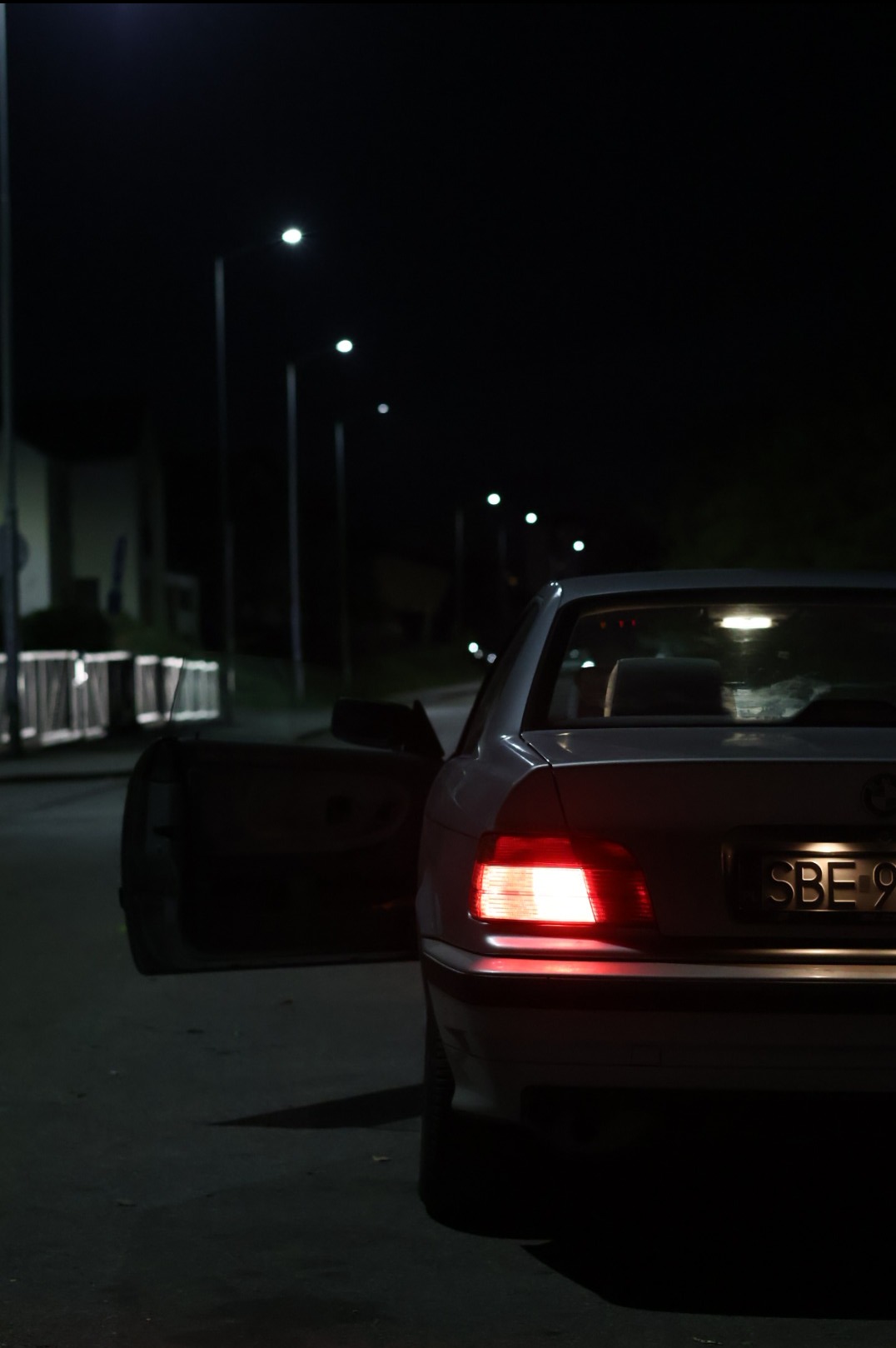 Tył zaparkowanego samochodu BMW z otwartymi drzwiami w nocy, oświetlony światłami ulicznymi i tylnymi światłami samochodu.