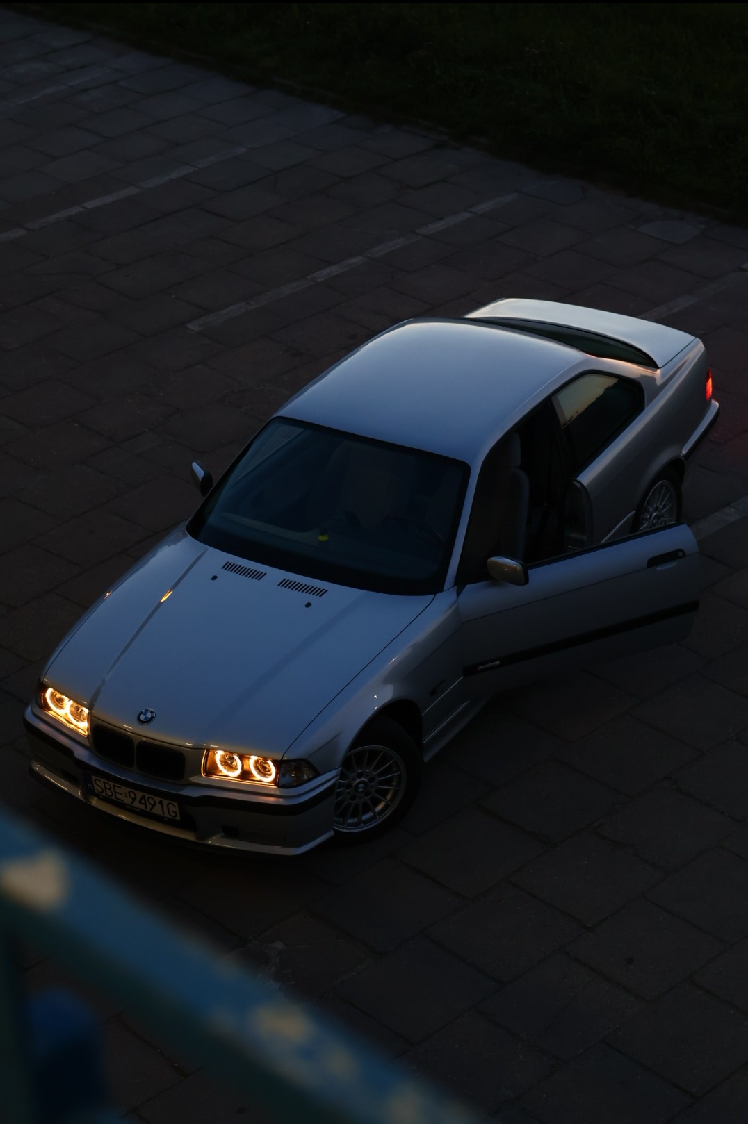 Srebrne BMW E36 coupe z otwartymi drzwiami kierowcy, zaparkowane na brukowanym parkingu o zmierzchu, z charakterystycznymi ringami świetlnymi w reflektorach.