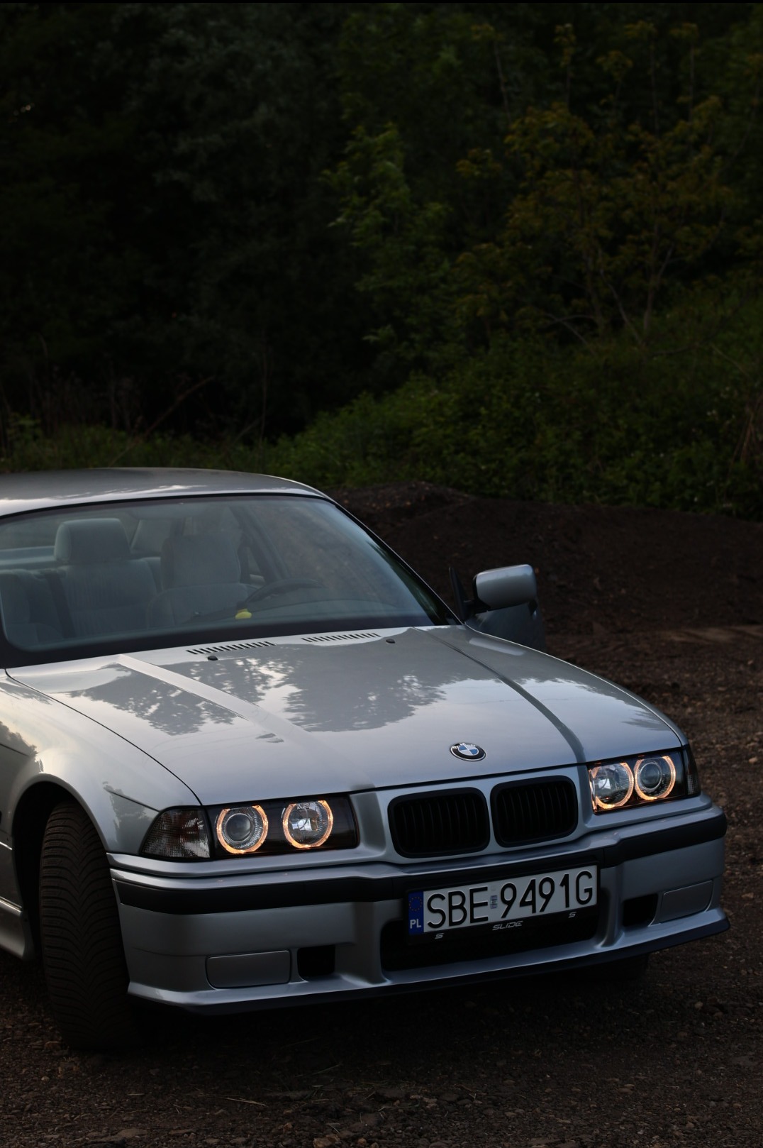 Srebrny samochód BMW E36 z charakterystycznymi ringami świetlnymi w reflektorach, zaparkowany na ziemi z tłem z zieleni.