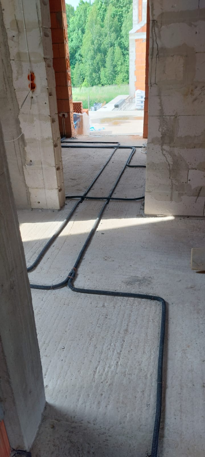 Wnętrze w stanie surowym z widocznymi peszlami instalacji elektrycznej ułożonymi na podłodze, w tle widok na zieleń przez niezamknięty otwór okienny.