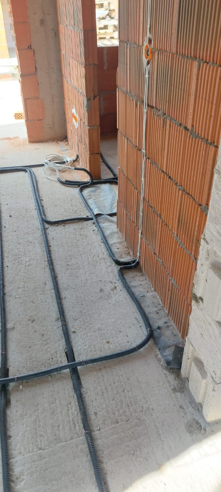 Instalacja przewodów elektrycznych w peszlach, ułożonych na podłodze w surowym stanie, przy ścianie z cegły dziurawki z zamontowaną puszką elektryczną.