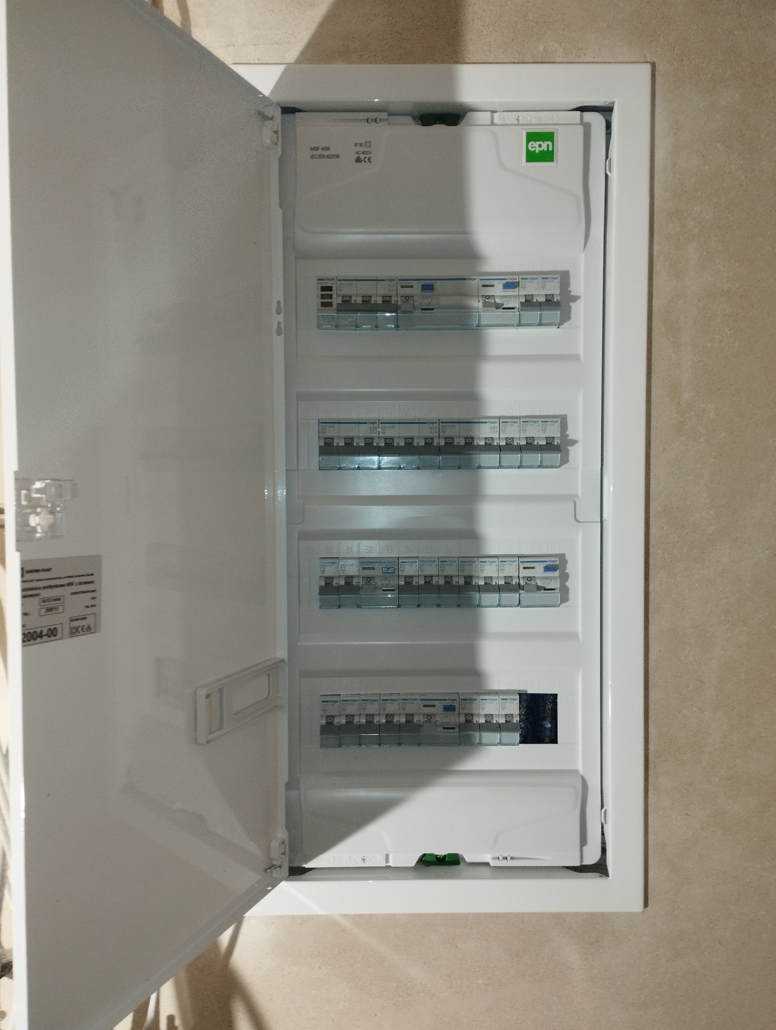 Otwarta, biała skrzynka elektryczna z bezpiecznikami Hager na beżowej ścianie. Widoczne oznaczenia MSF 456 i logo EPN. Instalacja wewnątrz budynku.