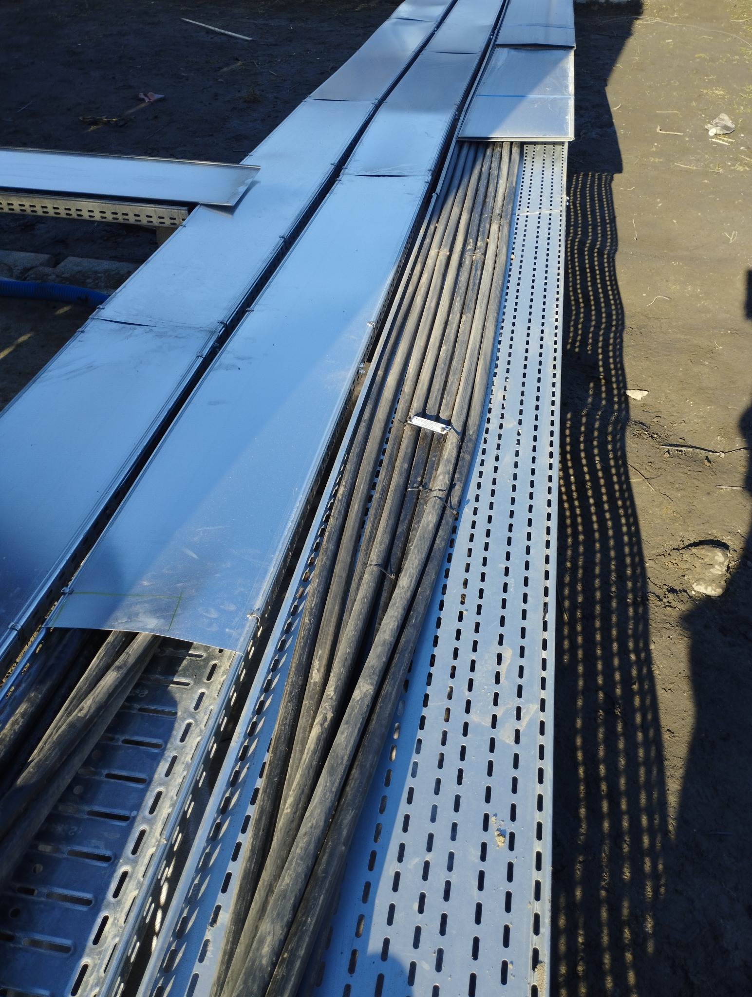 Metalowe korytka kablowe z ułożonymi przewodami, widoczne perforacje i błyszcząca powierzchnia, cień osoby w prawym rogu.