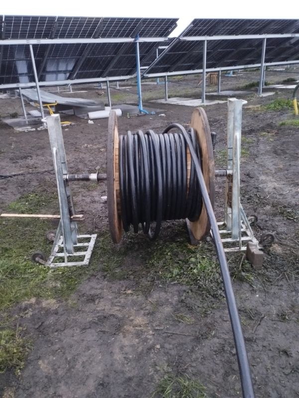 Czarny kabel na drewnianej szpuli z metalowym stojakiem na błotnistym terenie, w tle panele słoneczne. Widok z dołu.