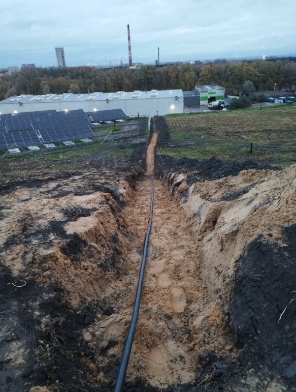 Czarny kabel elektryczny ułożony w wykopie ziemnym, w tle panele słoneczne i budynki przemysłowe. Widok z góry na instalację przyłącza elektrycznego.