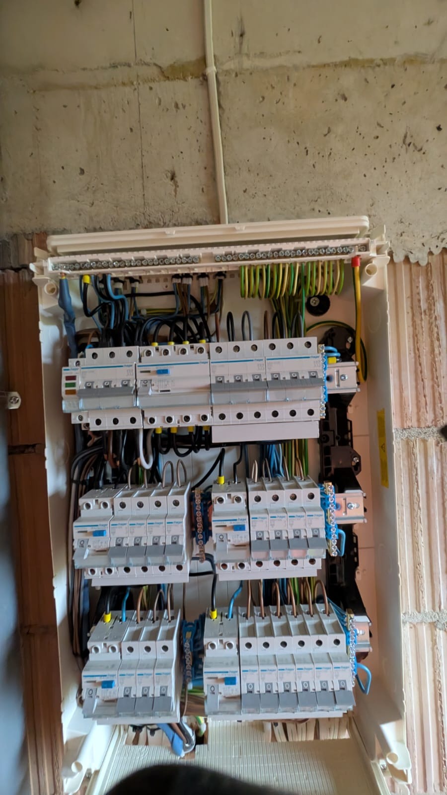 Rozdzielnica elektryczna z zamontowanymi bezpiecznikami i okablowaniem, widoczna na tle surowej ściany. Górna część rozdzielnicy z listwą zaciskową.