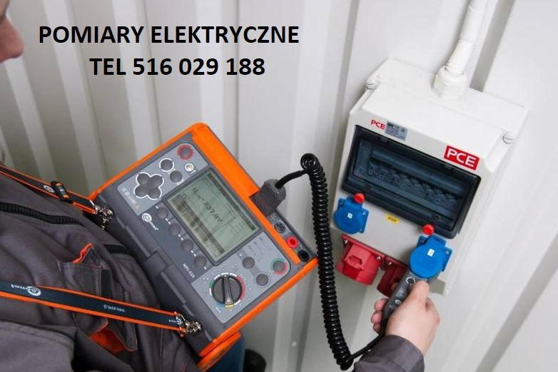 Elektryk w szarej kurtce, z miernikiem w pomarańczowej obudowie zawieszonym na szyi, sprawdza napięcie w gniazdach elektrycznych w białej skrzynce bezpiecznikowej PCE na tle białej ściany.