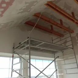 Sufit poddasza w trakcie prac wykończeniowych, widoczne drewniane elementy konstrukcyjne, częściowo zagruntowany sufit oraz rusztowanie na tle okna.