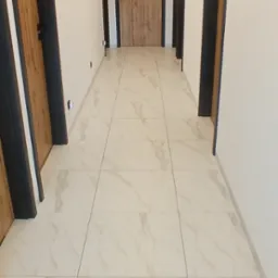 Długi korytarz z białymi ścianami, podłogą wyłożoną białymi płytkami imitującymi marmur oraz drewnianymi drzwiami z czarnymi ościeżnicami i klamkami, oświetlony listwą z punktowymi lampami sufitowymi.