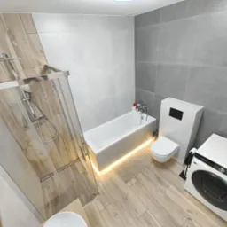 Nowoczesna łazienka z drewnopodobnymi płytkami, kabiną prysznicową typu walk-in, podświetlaną wanną, podwieszaną toaletą i pralką Samsung, prezentująca minimalistyczny design.