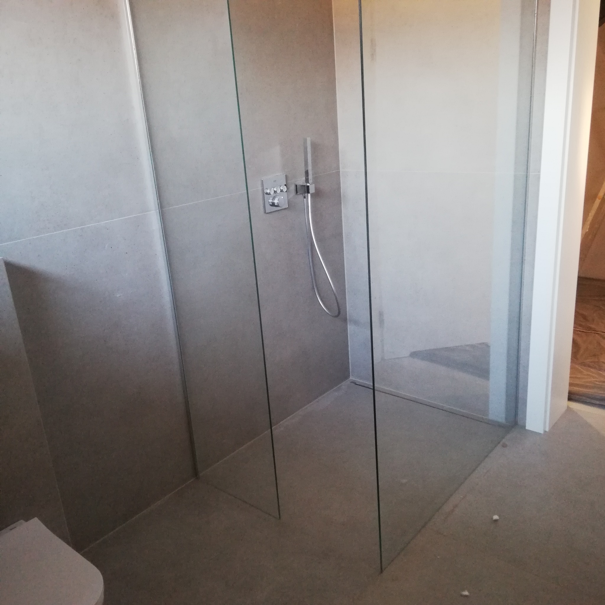 Nowoczesna kabina prysznicowa walk-in z szarymi płytkami, minimalistyczna armatura i częściowo widoczna toaleta w rogu.