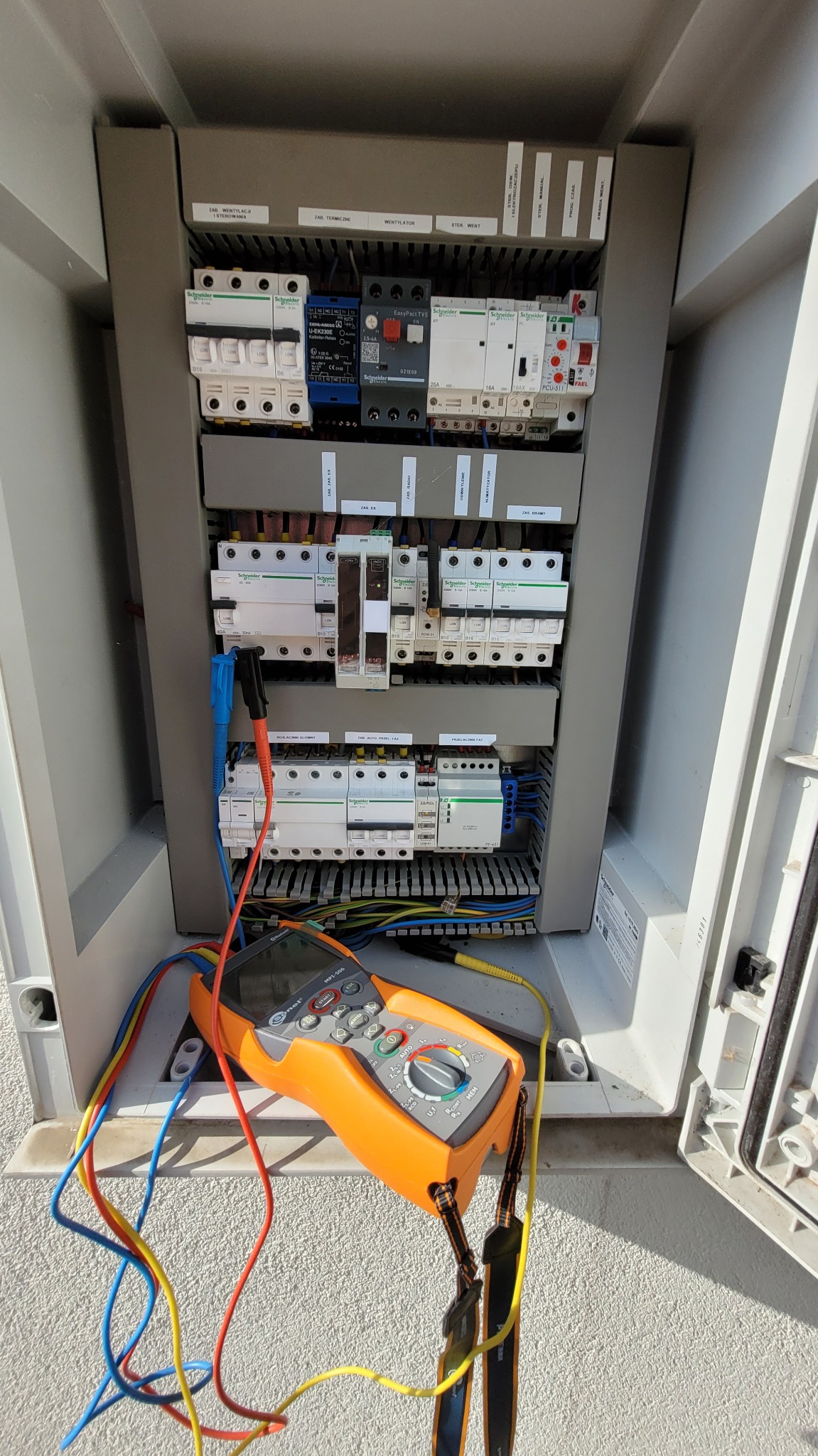 Panel elektryczny z multimetrem w Warszawie. Widoczne bezpieczniki, kable i urządzenie pomiarowe. Skrzynka sterownicza z elementami automatyki.