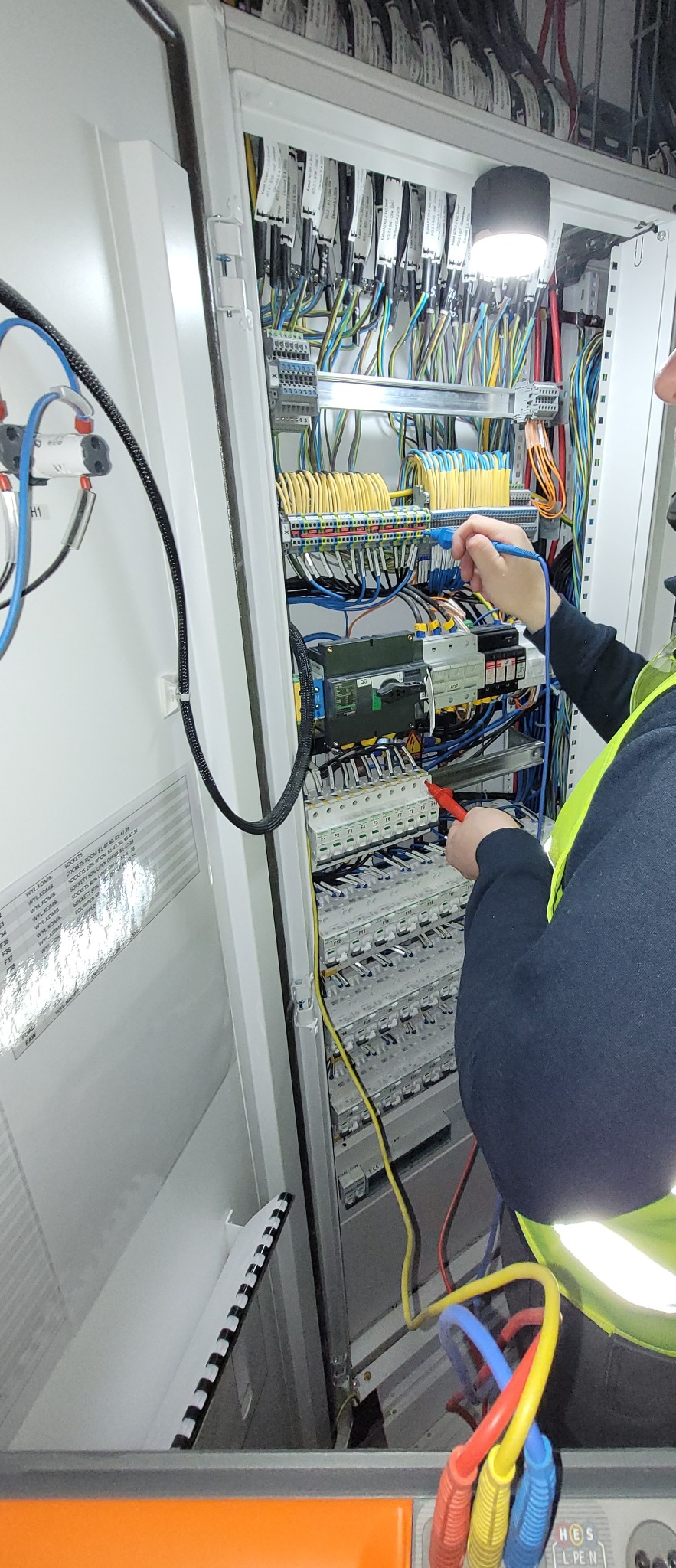 Elektryk w Warszawie, w kamizelce odblaskowej, mierzy napięcie w rozdzielnicy elektrycznej za pomocą miernika; widoczne liczne przewody, listwy zaciskowe i dokumentacja technicza.