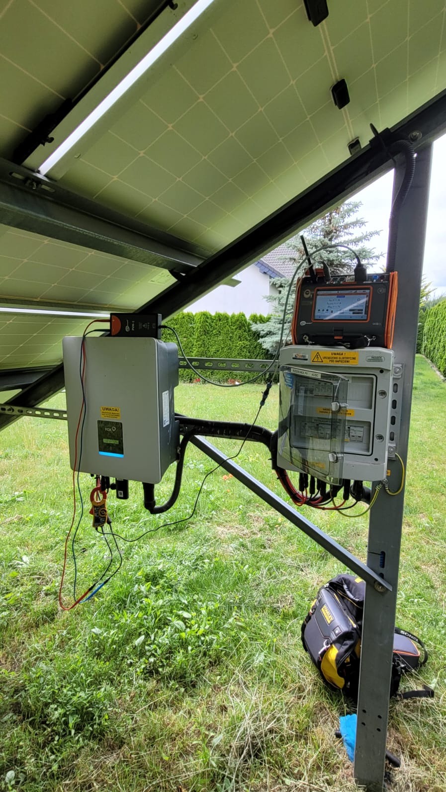 Instalacja fotowoltaiczna: Inwerter, skrzynka z zabezpieczeniami i miernik na tle zieleni. Widoczny też częściowo panel słoneczny i torba narzędziowa.