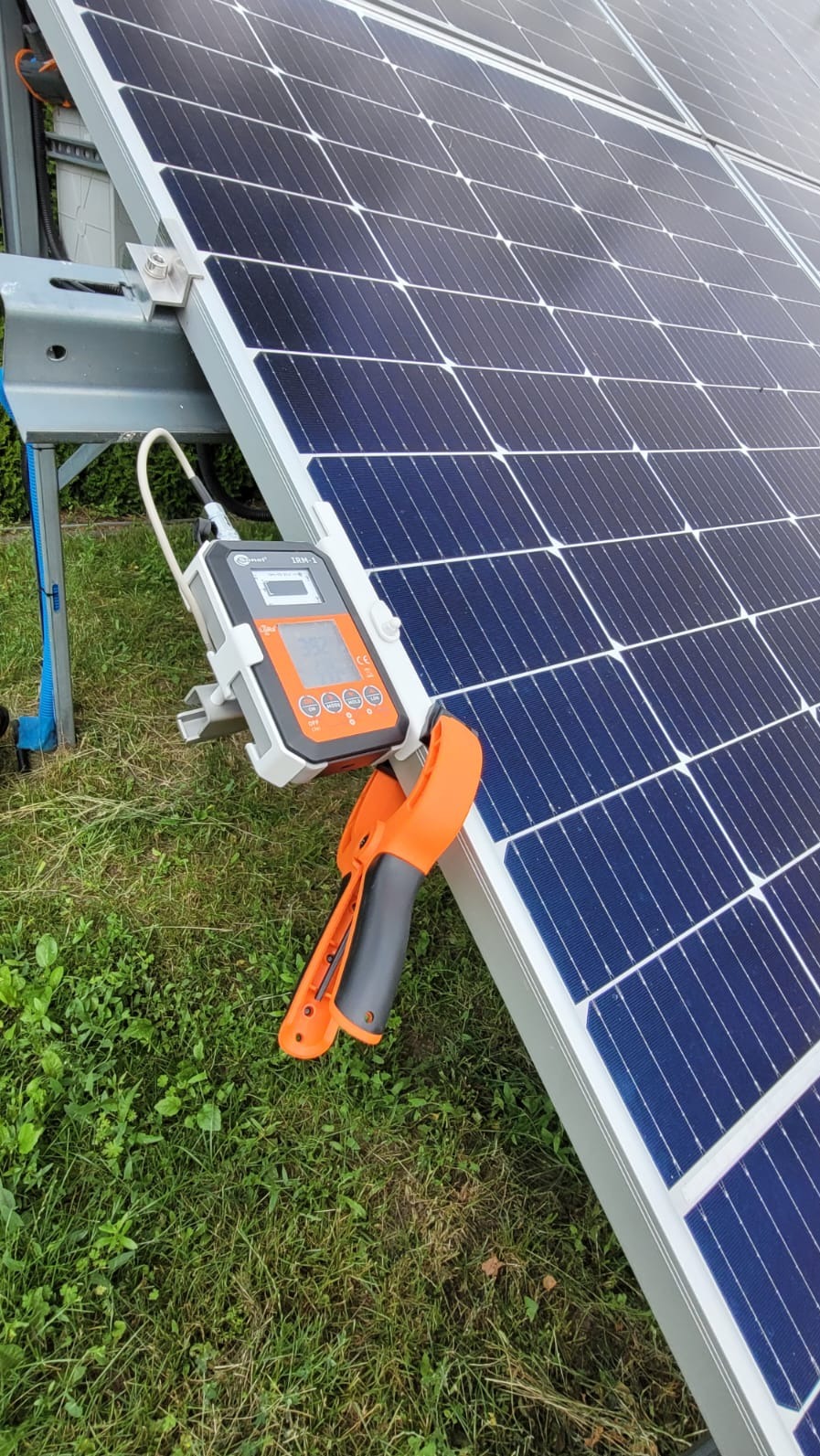 Panel słoneczny z zamocowanym miernikiem elektrycznym i cęgami, na tle trawy. Pomiar napięcia instalacji fotowoltaicznej.