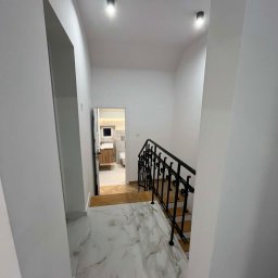 MULTIMAKE SPÓŁKA Z OGRANICZONĄ ODPOWIEDZIALNOŚCIĄ - Schody z marmurowymi płytkami i czarną, kutą balustradą. W tle widoczne wejście do łazienki z drewnianą szafką i białym wyposażeniem. Minimalistyczne oświetlenie.