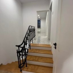 MULTIMAKE SPÓŁKA Z OGRANICZONĄ ODPOWIEDZIALNOŚCIĄ - Nowoczesne schody z drewnianymi stopniami i czarną, kutą balustradą w jasnym wnętrzu. Widoczny fragment parkietu i drzwi.