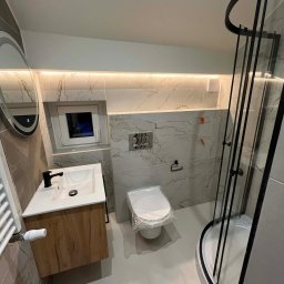 MULTIMAKE SPÓŁKA Z OGRANICZONĄ ODPOWIEDZIALNOŚCIĄ - Nowoczesna łazienka po remoncie: kabina prysznicowa z czarnymi ramami, umywalka z drewnianą szafką, marmurowe płytki i podświetlane lustro. Toaleta zabezpieczona folią.