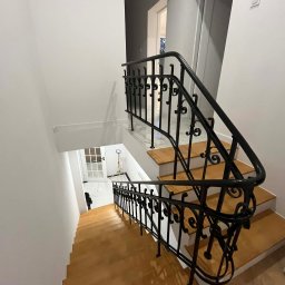 MULTIMAKE SPÓŁKA Z OGRANICZONĄ ODPOWIEDZIALNOŚCIĄ - Klatka schodowa z drewnianymi stopniami i ozdobną, czarną, kutą balustradą. Widok z góry na dół, białe ściany, drzwi w tle. Charakterystyczny wzór balustrady.