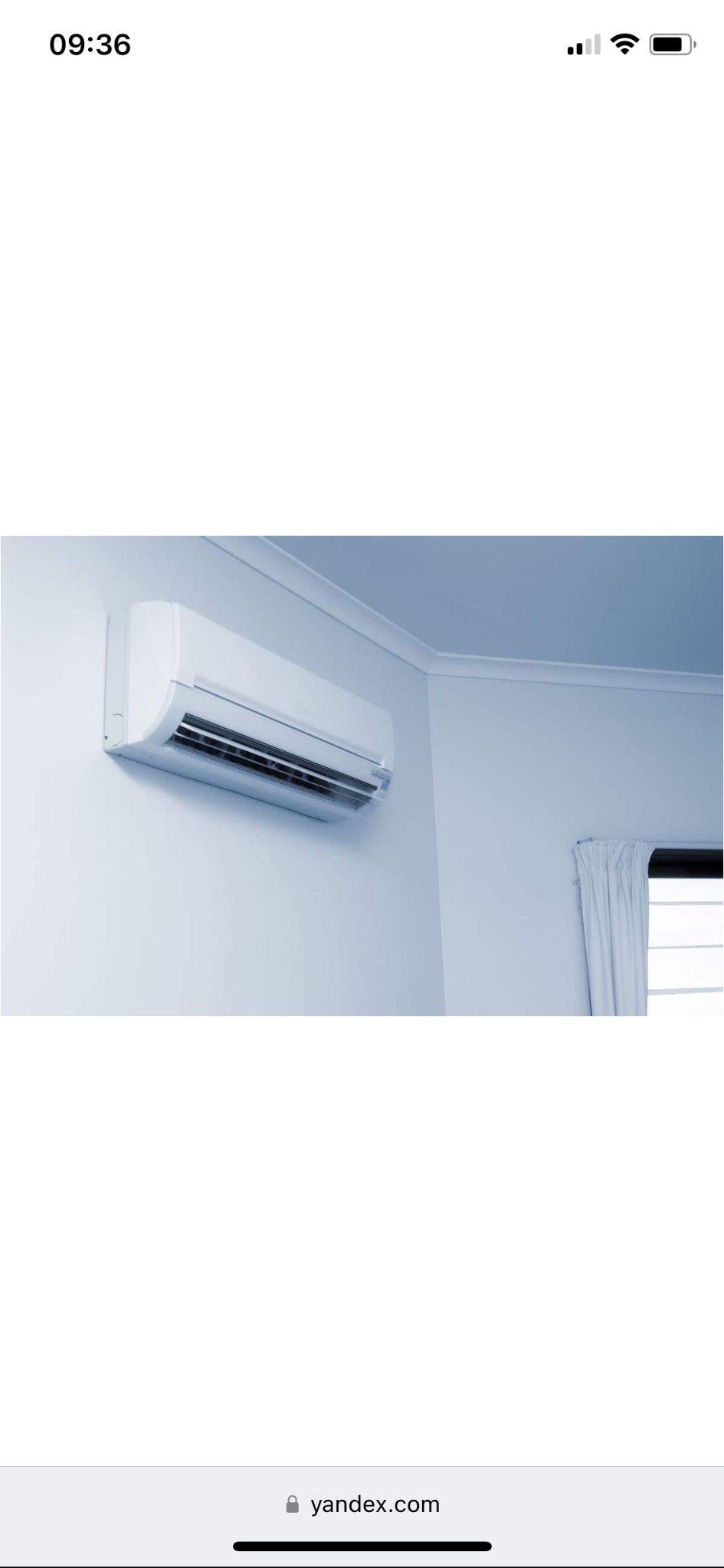 Biały klimatyzator ścienny zamontowany w rogu pokoju, widoczne białe ściany, sufit z listwami oraz okno z zasłoną.