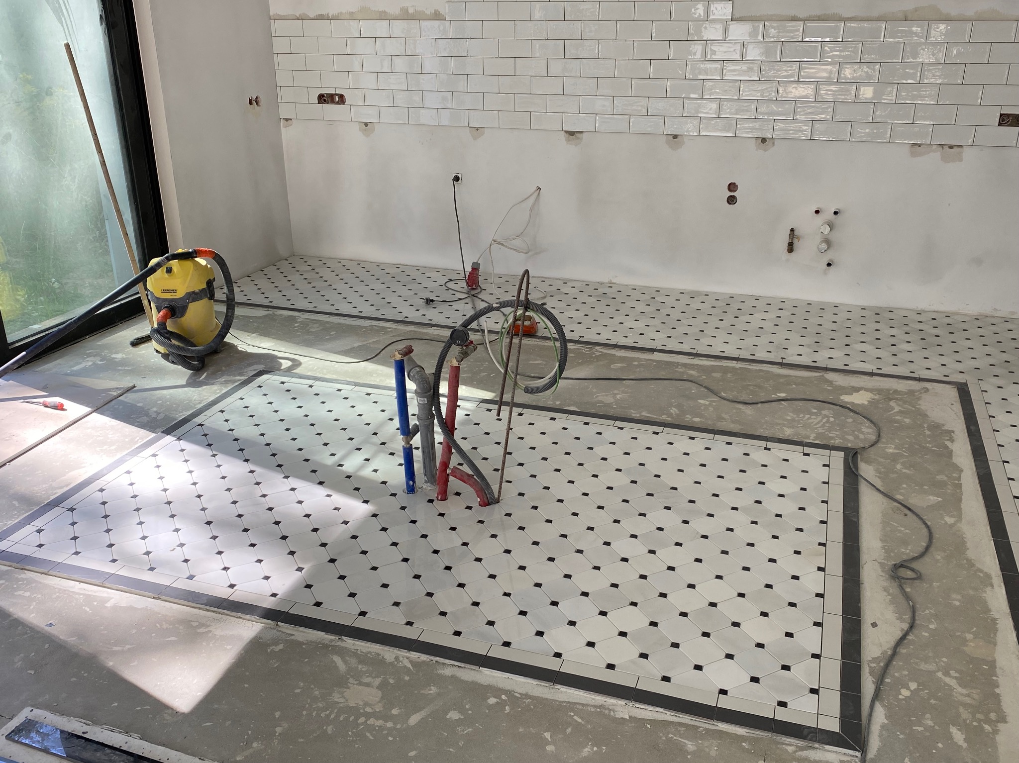 Ukończona podłoga z białych płytek z czarnymi akcentami i obramowaniem. Częściowo wykończona ściana z białych płytek. Widoczny odkurzacz budowlany i rury.