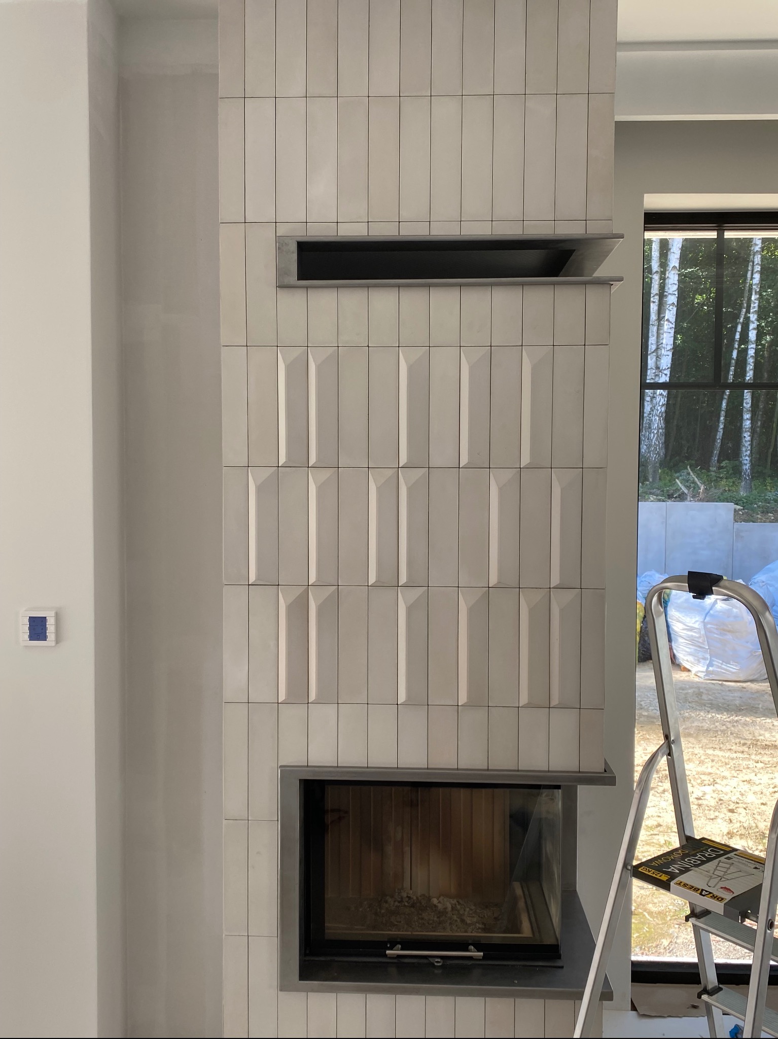 Wykończony kominek z jasnymi, pionowo ułożonymi płytkami o geometrycznym wzorze. Drabina oparta o ścianę obok okna z widokiem na zewnątrz.