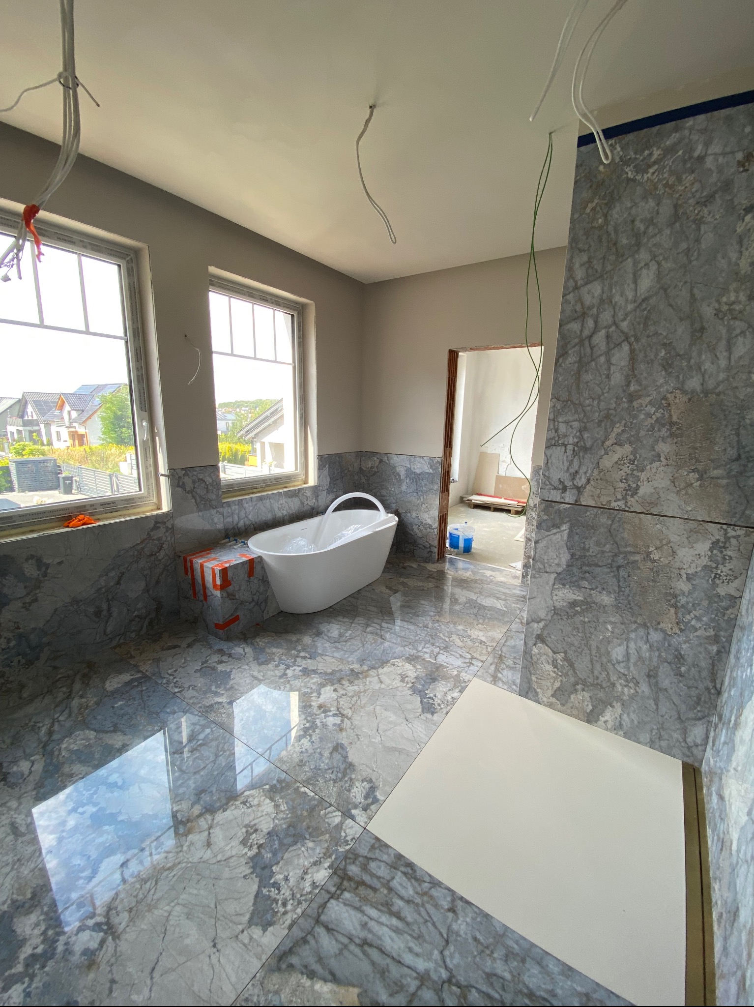 Łazienka w trakcie wykończenia: marmurowe płytki, wolnostojąca wanna, okna z widokiem na domy, widoczne przewody instalacji elektrycznej.