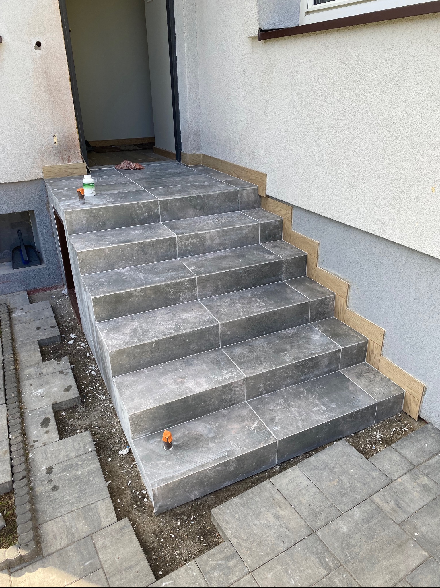 Wyłożone szarymi płytkami schody wejściowe do budynku, z widocznymi elementami montażowymi i częściowo ułożoną kostką brukową. Jasna elewacja budynku w tle.