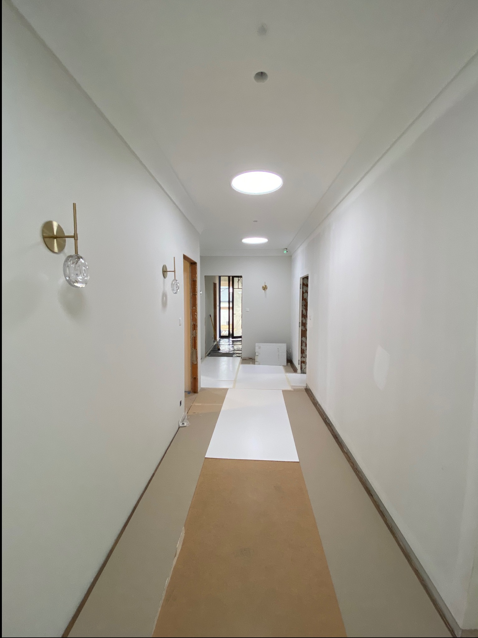 Długi, biały korytarz w trakcie wykańczania z dekoracyjnymi lampami ściennymi i zabezpieczoną podłogą. Widoczne otwory w suficie.