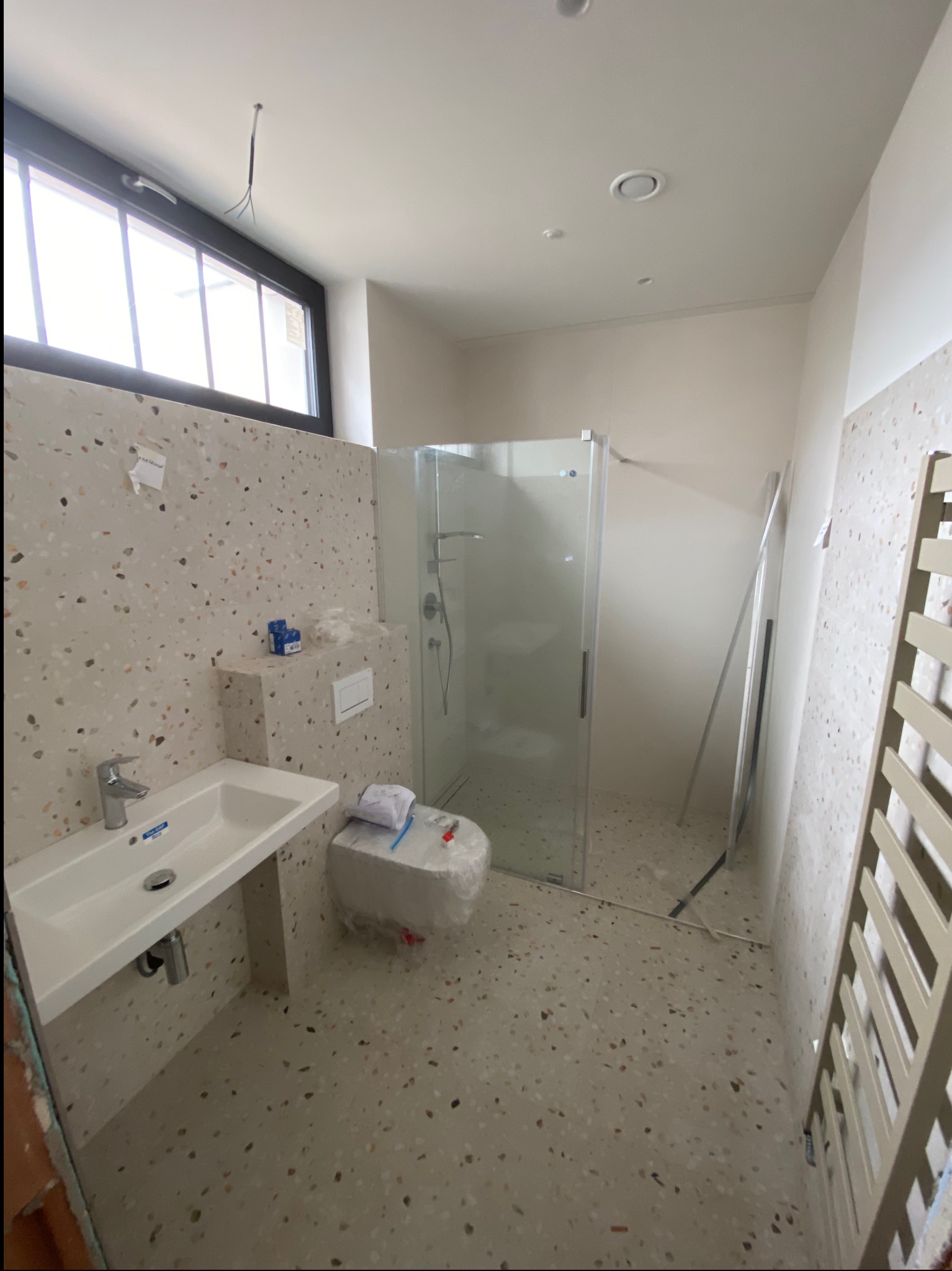 Remont łazienki: Umywalka, toaleta w folii, kabina prysznicowa i grzejnik na tle ścian i podłogi z lastryko. Widoczne przewody elektryczne.