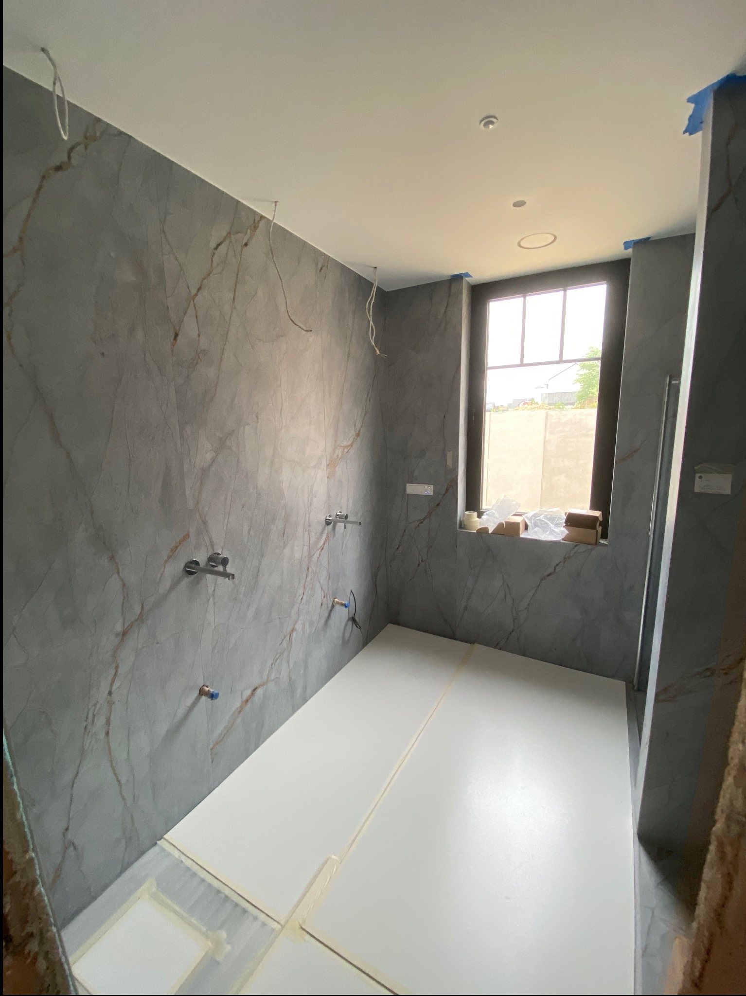 Łazienka w trakcie remontu: szare płytki imitujące marmur na ścianach, biała podłoga, okno z widokiem na zewnątrz. Widoczne instalacje wodne i pozostałości po pracach.