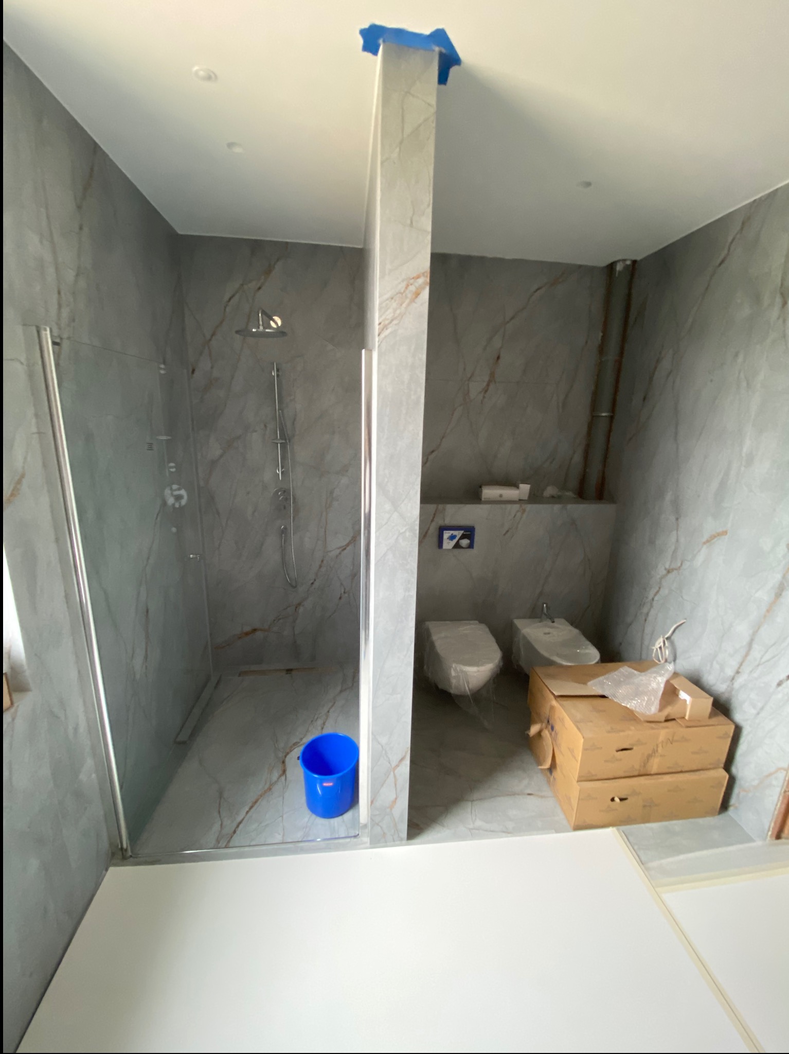 Wnętrze łazienki w trakcie wykończenia z kabiną prysznicową, toaletą i bidetem, wykończone marmurowymi płytkami. Widoczne kartony i niebieskie wiadro.