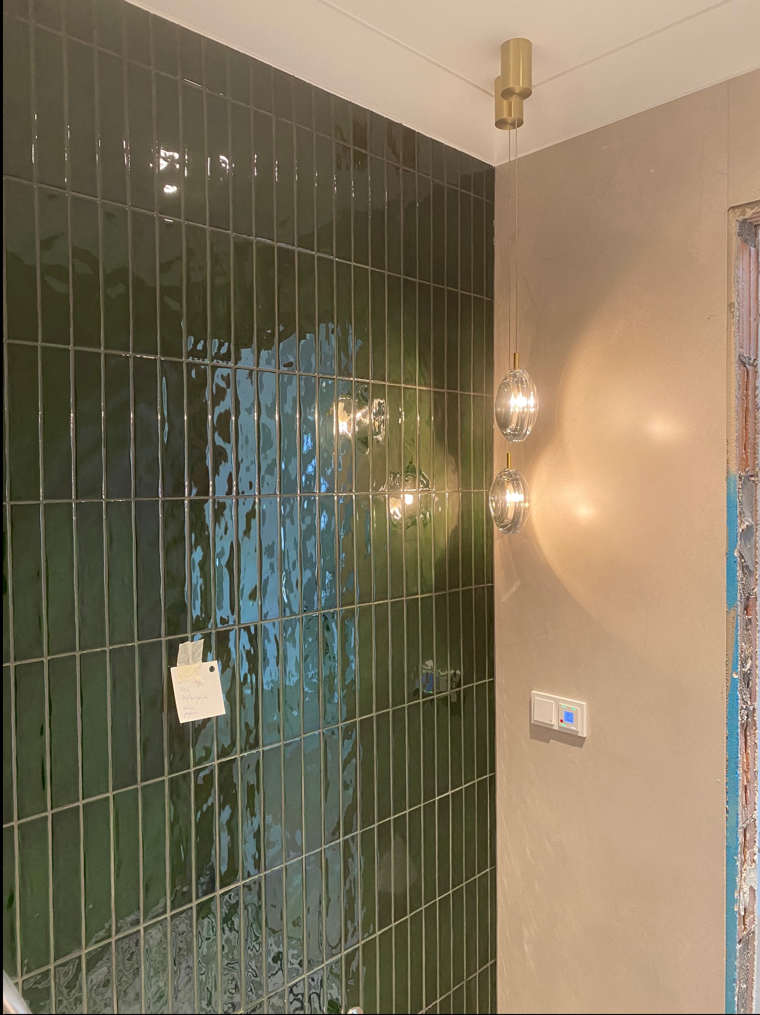 Ściana wyłożona pionowymi, błyszczącymi, butelkowozielonymi płytkami, oświetlona wiszącą lampą z dwoma szklanymi kloszami. Widoczny fragment ściany z gładzią.