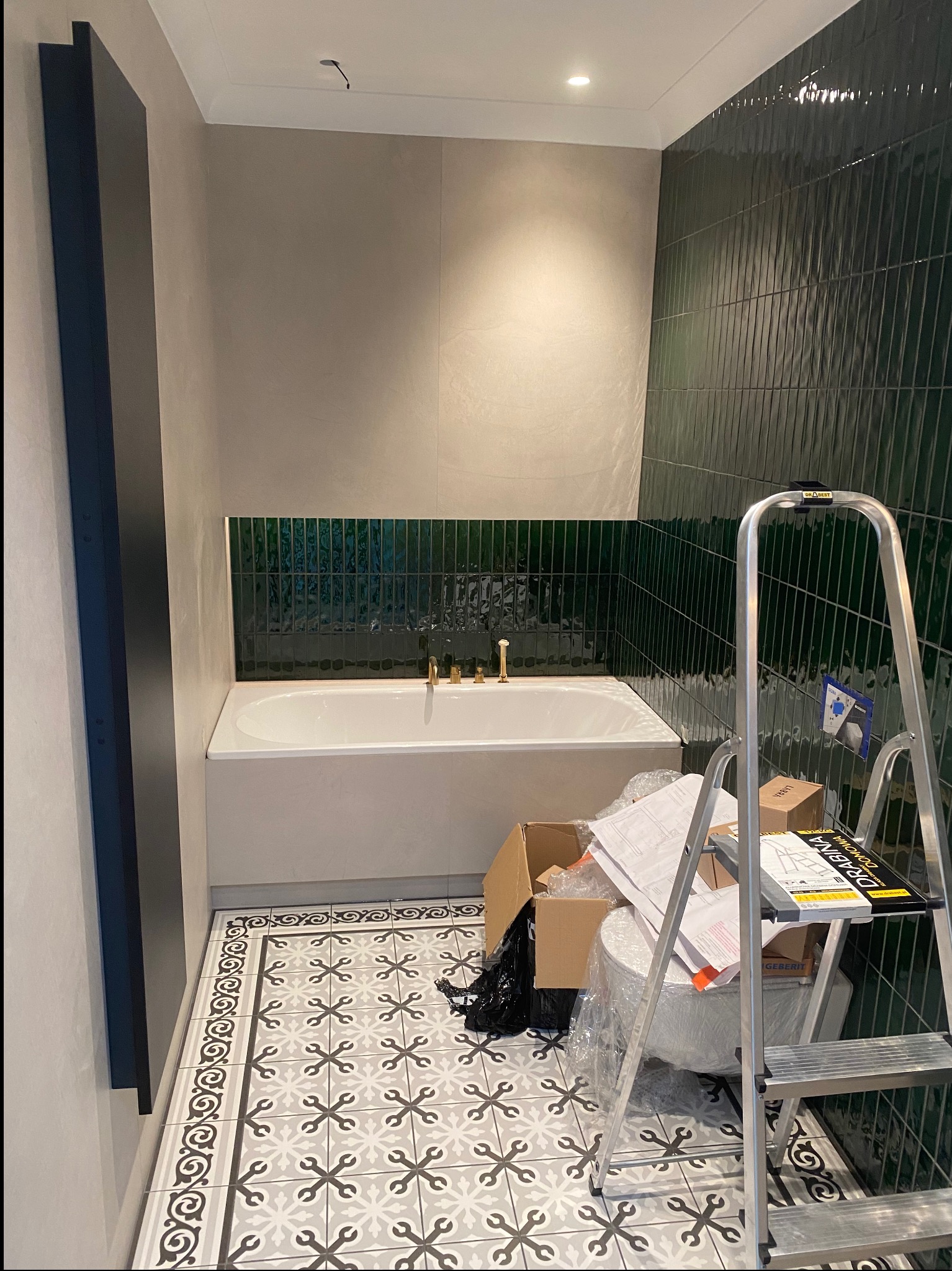 Remont łazienki: biała wanna z złotą armaturą, ściana z ciemnozielonych płytek, dekoracyjna podłoga. Aluminiowa drabina stoi obok toalety.