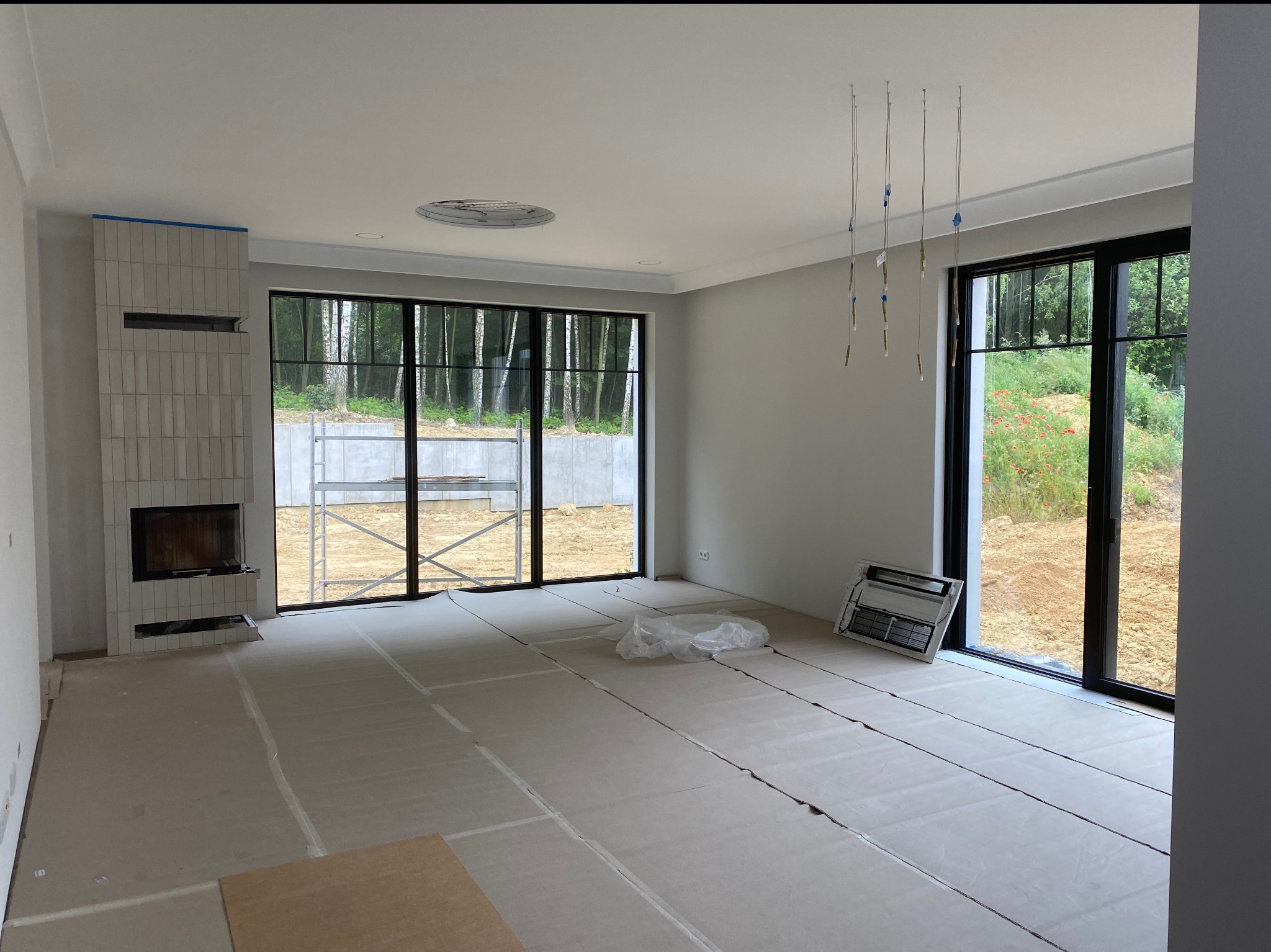 Wnętrze w trakcie wykończenia: zabezpieczona podłoga, obudowany kominek z płytek, duże okna z widokiem na zewnątrz. Widoczne elementy instalacji oświetleniowej.
