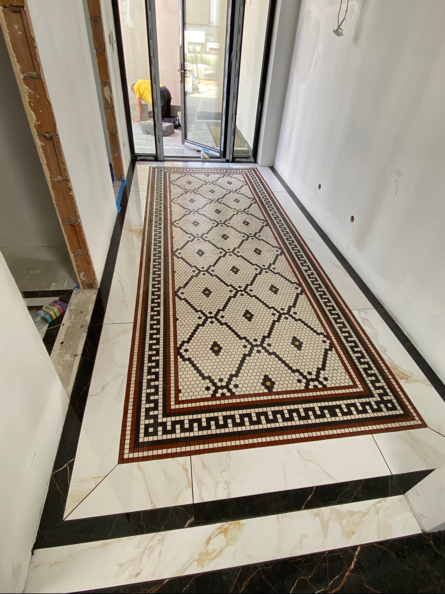 Ukończona podłoga w korytarzu z mozaiką w stylu retro, obramowana marmurowymi płytkami. W tle widoczna osoba pracująca przy wejściu.