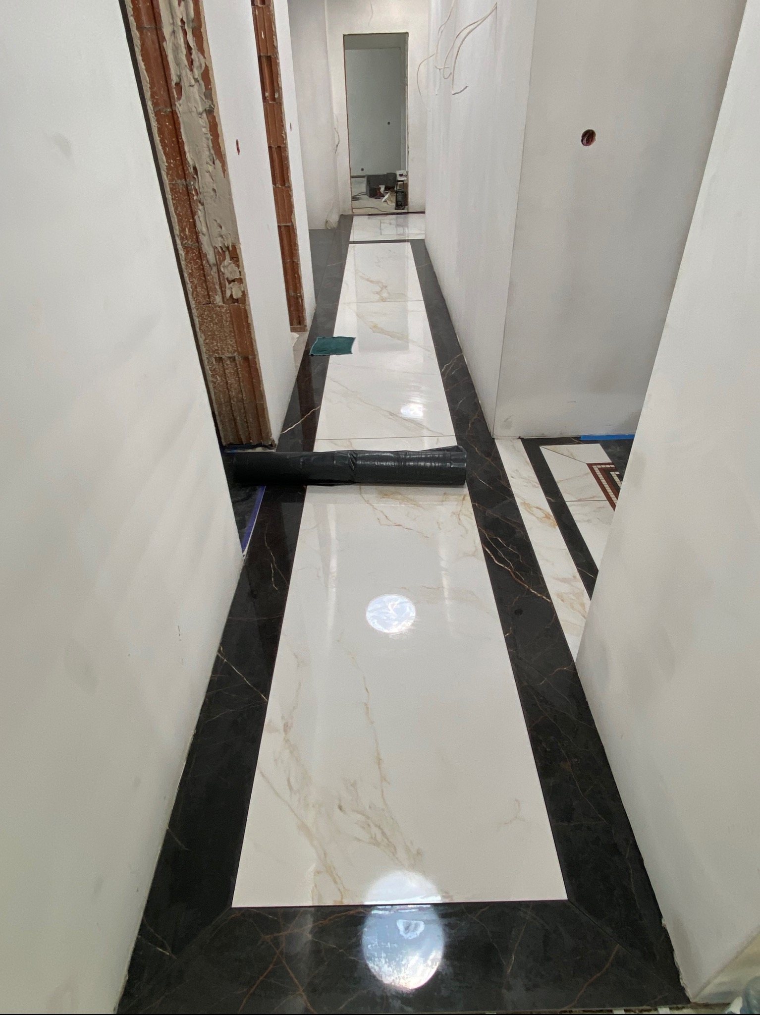Wnętrze korytarza w trakcie wykańczania: ułożona, błyszcząca podłoga z białych i czarnych płytek imitujących marmur, ściany przygotowane do malowania, widoczne kable.