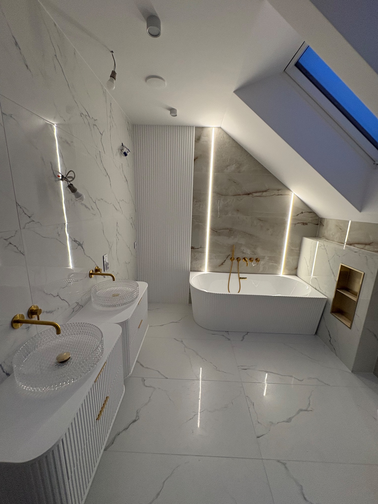 Elegancka łazienka na poddaszu z marmurowymi płytkami, wolnostojącą wanną i szklanymi umywalkami na białych szafkach z ryflowanymi frontami. Złote baterie i oświetlenie LED.