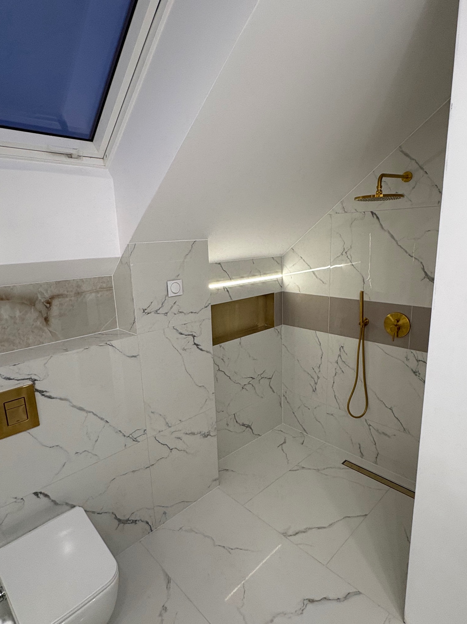 Wykończona łazienka na poddaszu z marmurowymi płytkami, złotą armaturą i oknem dachowym. Uwagę przykuwa minimalistyczny design i oświetlenie LED w półce.