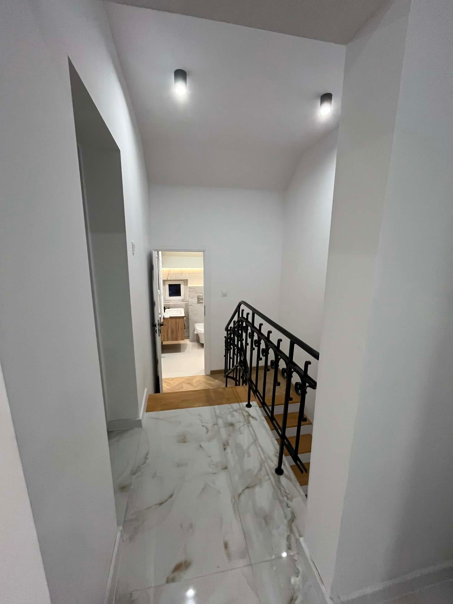 Schody z marmurowymi płytkami i czarną, kutą balustradą. W tle widoczne wejście do łazienki z drewnianą szafką i białym wyposażeniem. Minimalistyczne oświetlenie.