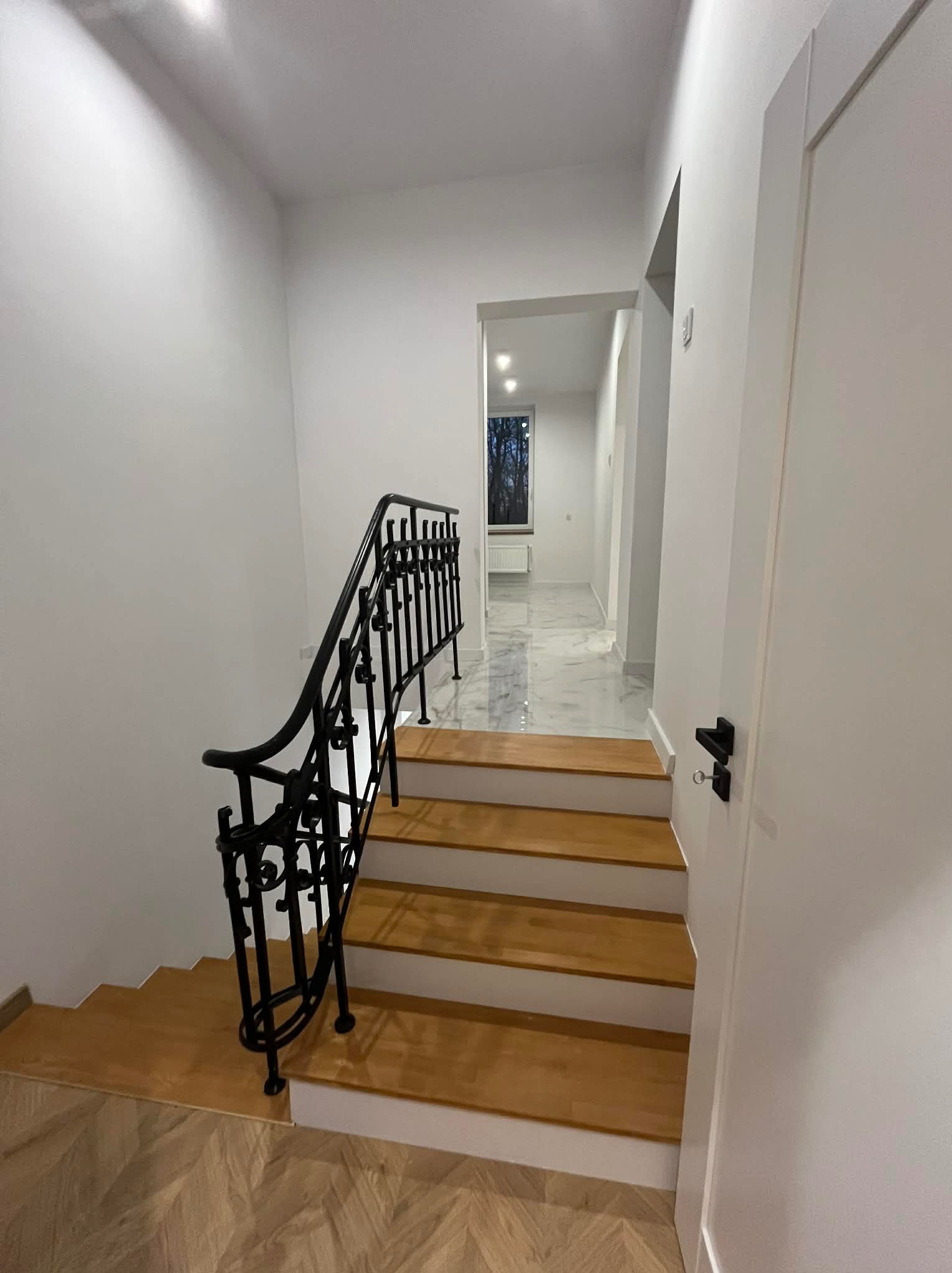 Nowoczesne schody z drewnianymi stopniami i czarną, kutą balustradą w jasnym wnętrzu. Widoczny fragment parkietu i drzwi.